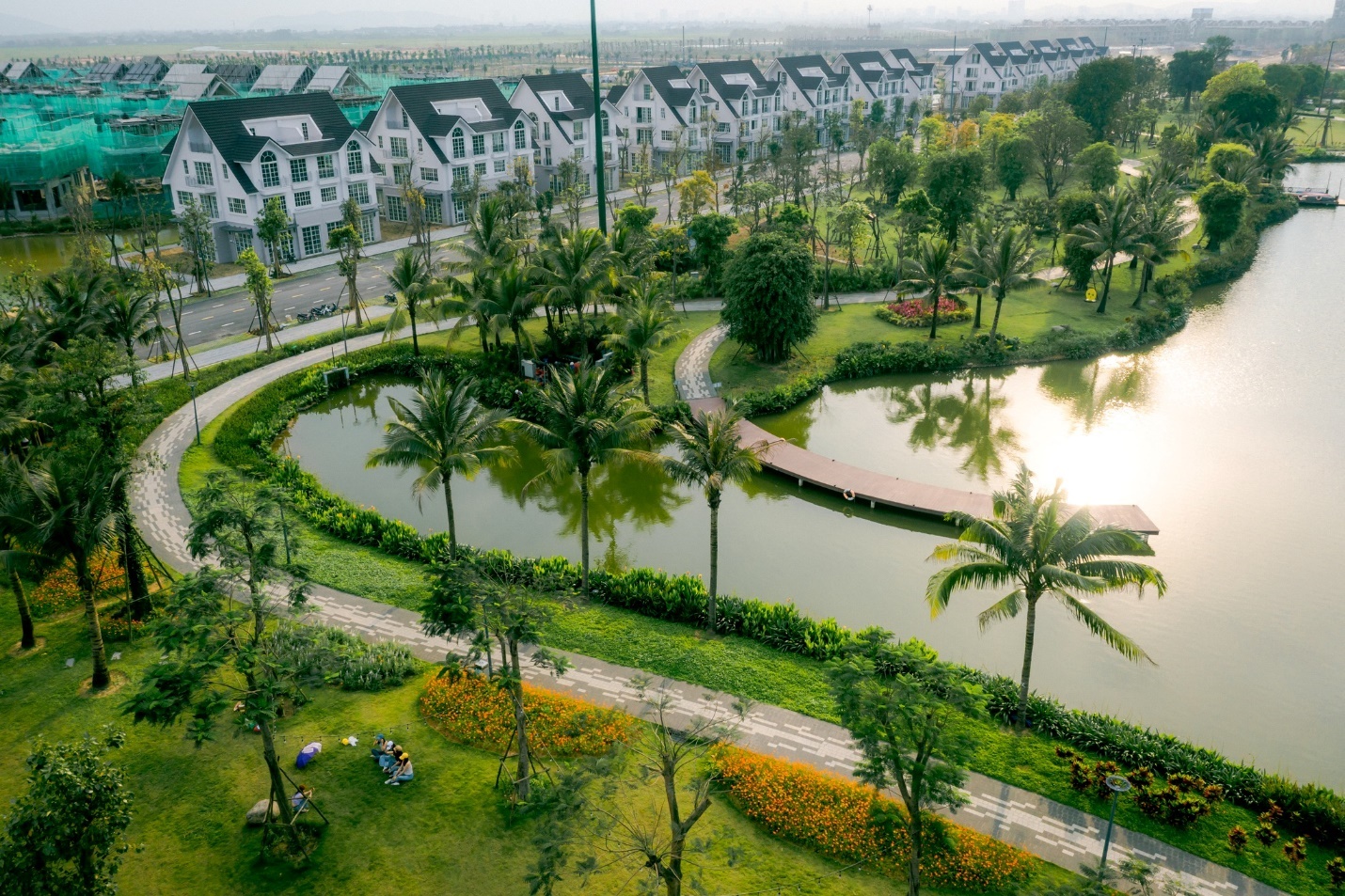 Giải phóng đôi chân, trải nghiệm mùa hè rực rỡ tại “Lễ hội mùa hè Eco Central Park 2025”- Ảnh 7. A pond with trees and a path in front of a row of buildings Description automatically generated