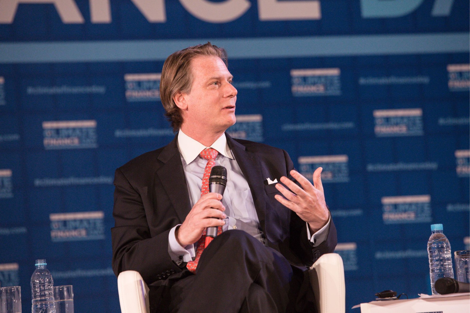 A person in a suit and tie speaking into a microphone  Description automatically generated