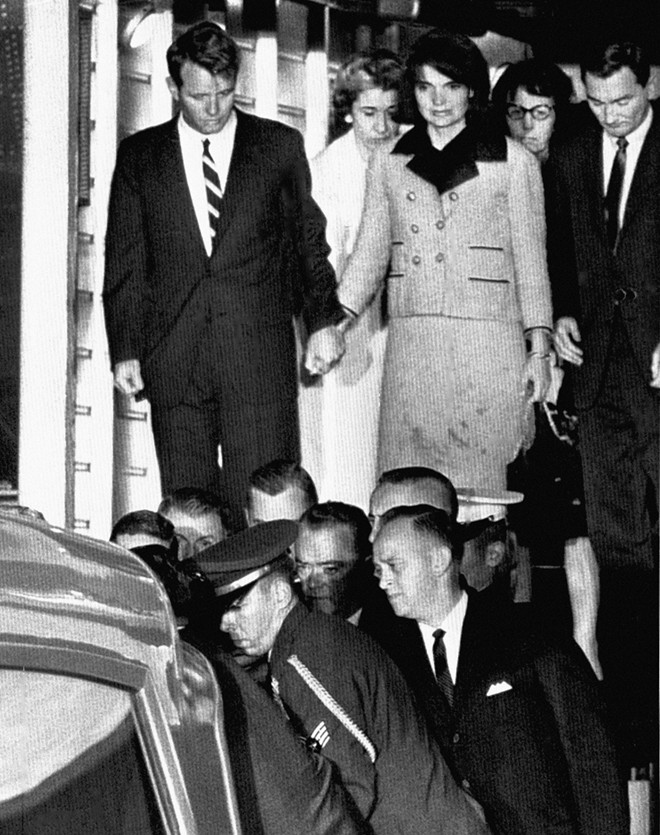 Phu nhân Jacqueline mặc bộ đồ còn nguyên những vết máu vấy từ thi thể chồng, nắm tay anh là Bộ trưởng Tư pháp Robert Kennedy. Trong khi đó, lực lượng an ninh nhấc quan tài đặt thi thể ông Kennedy từ một xe cứu thương. Lúc này, đoàn người đã đến căn cứ không quân Andrews gần Washington.